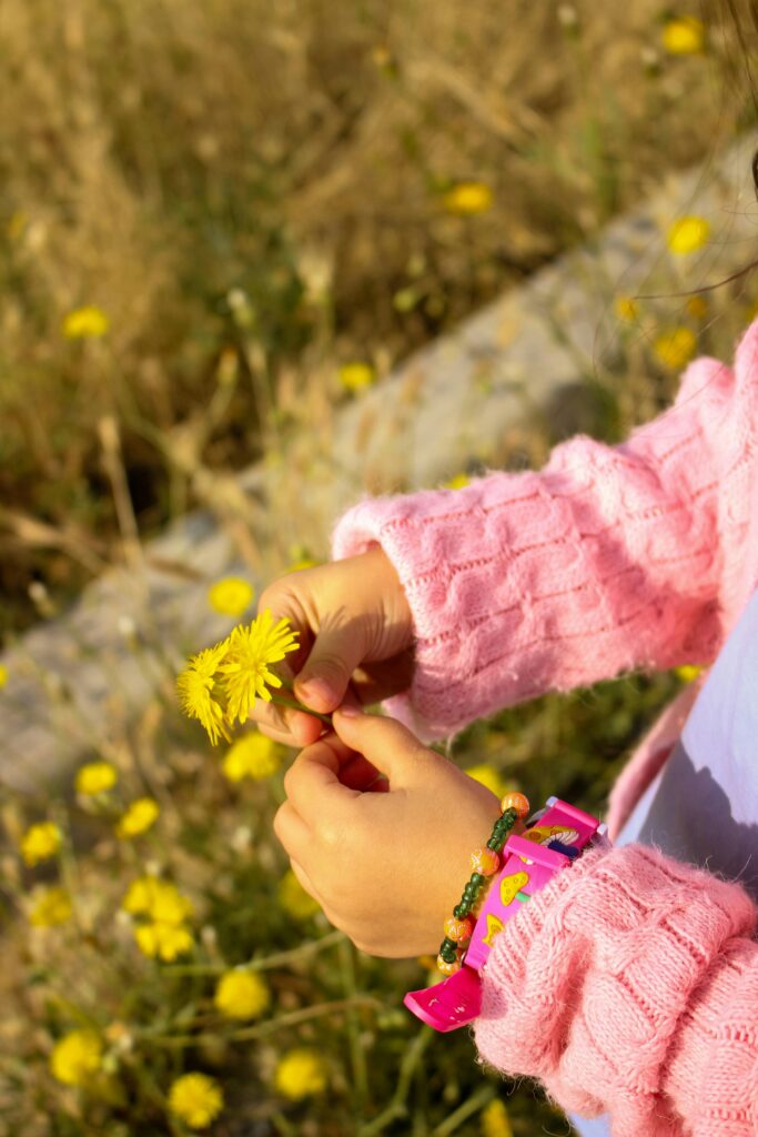 A child in a pink sweater holding vibrant yellow dandelions in a sunny meadow.